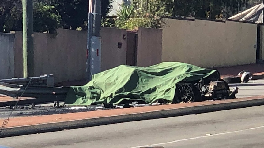The wreckage of a Ferrari lies under a green tarpaulin on a road.