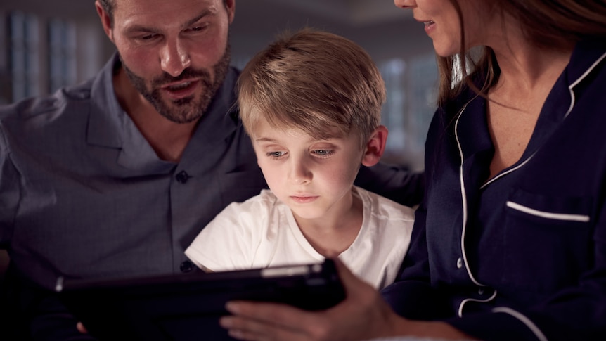 Parents using a tablet device with their young son in their living room.