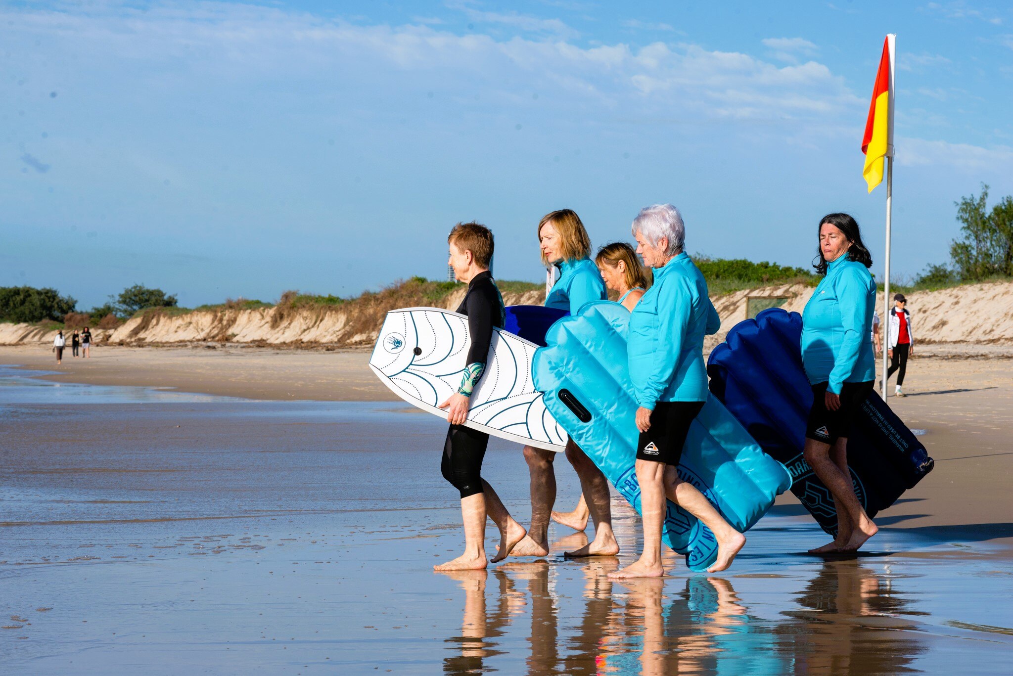 A group of older women with boogie boards under their arms walk into the water at a beach.