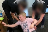 A 14-year-old boy screams, sitting on the floor as two men whose faces are blurred push him.