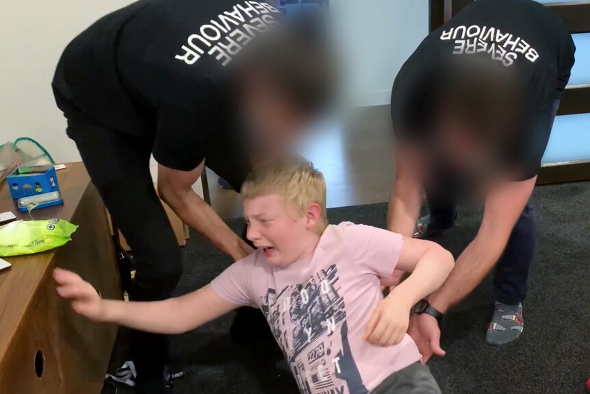 A 14-year-old boy screams, sitting on the floor as two men whose faces are blurred push him.