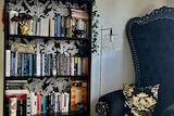 A living room corner with an ornate black chair on the right of a full bookcase lined with gothic wallpaper.