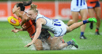 A woman tackles another woman to the ground as she tries to retain a football while water splashes in sodden conditions