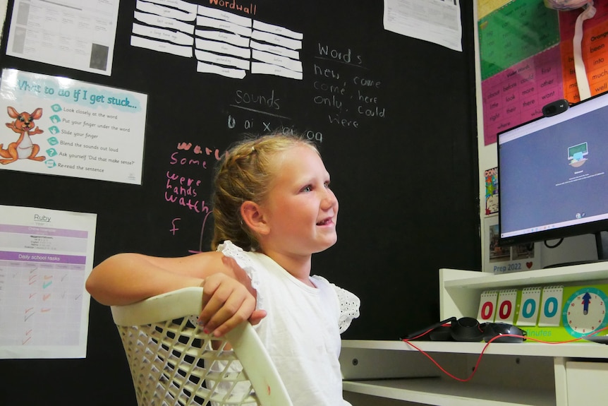 A young girl sits at a desk, behind her is a blackbaord wall with paper stuck to it and on the desk is a computer. 