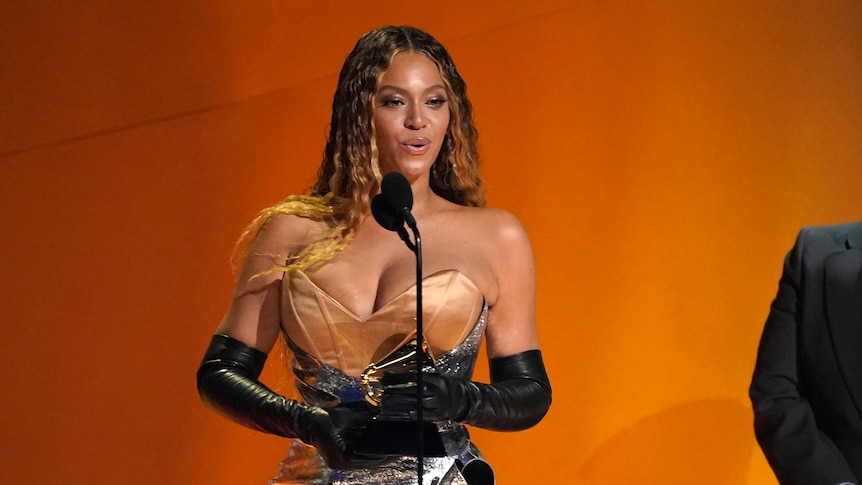 A good-looking black woman in a gold and silver dress holds a Grammys trophy onstage.