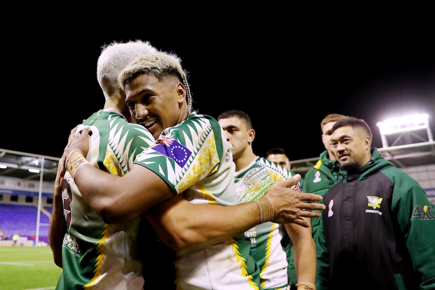 Two Cook Islands rugby league players hug after a match as their teammates watch on.