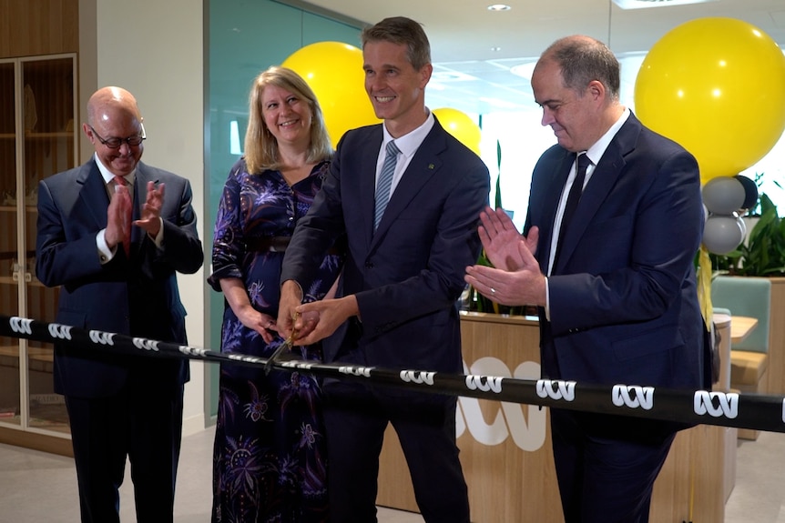 Three people in formal attire applaud as a man cuts a ribbon in an office. 