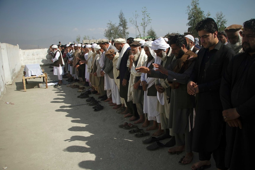 Dozens of men stand in a line with their eyes closed as they pray in front of a coffin, which is covered in a white sheet.