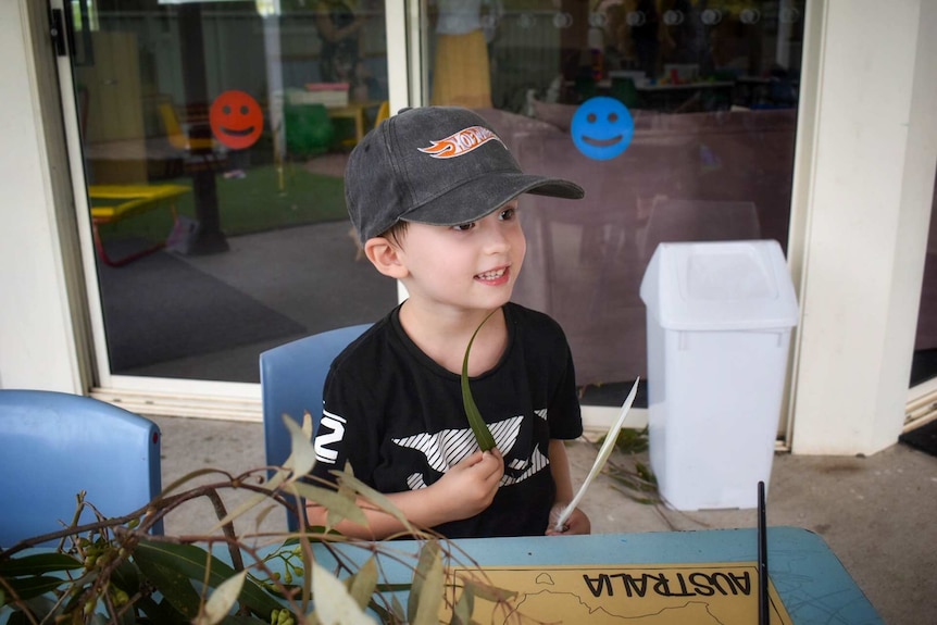Young boy with big smile sitting at desk outside with eucalyptus leaves in foreground.