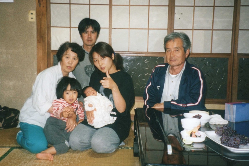 A woman kneeling on a tatami mat cradling a newborn, surrounded by her family