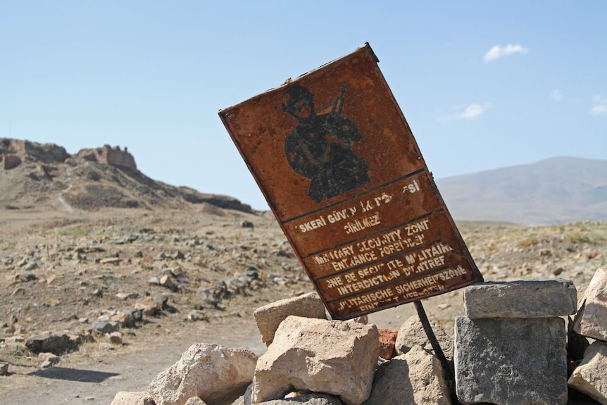 A rusty sign with warnings in various languages in the foreground of a barren, deserted landscape.