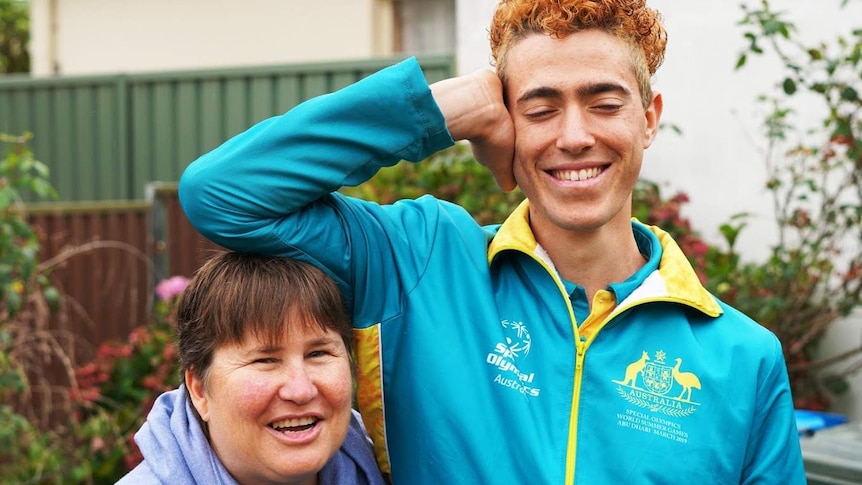 An older woman with short brown hair smiles at teh camera standing next to a tall young man resting his elbow on her head.