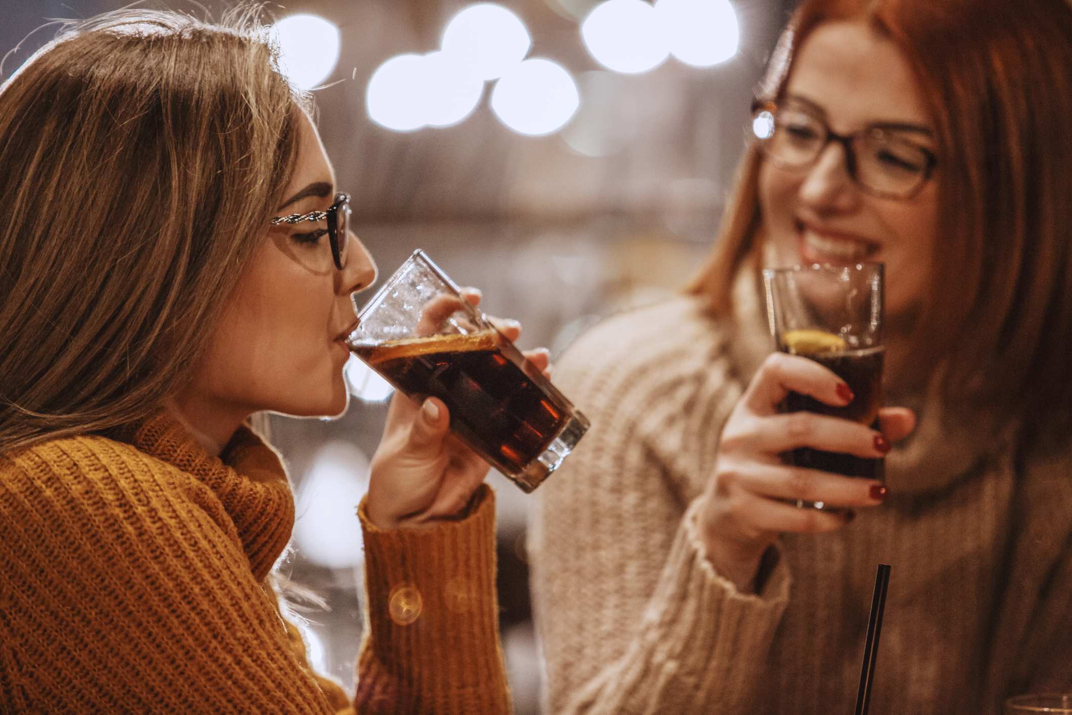 Two women smile as they drink cola with lemon wedges from glasses.