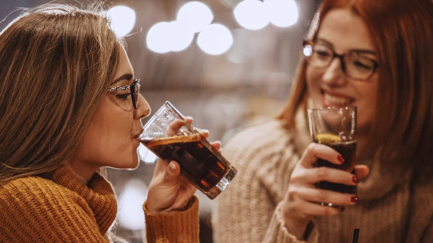 Two women smile as they drink cola with lemon wedges from glasses.