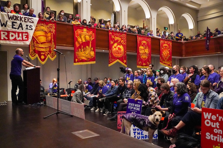 Dozens of union members holding signs fill the floors and internal balconies of Trades Hall, as a member speaks on stage.