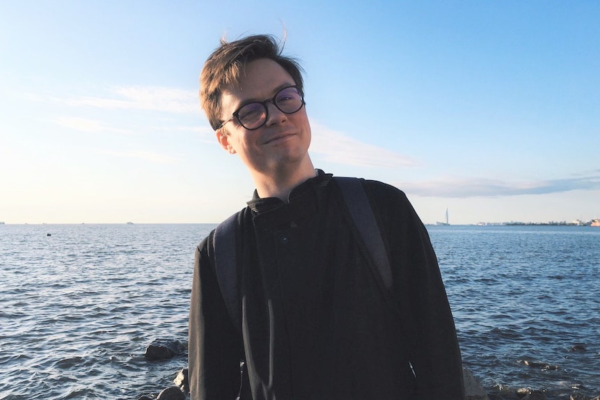 A man with brown hair and wearing glasses tilts his head as he stands infront of the ocean.