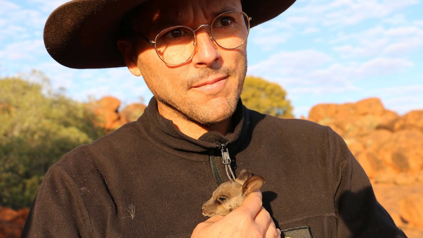 A man wearing glasses and a hat holding a very small wallaby close to his chest