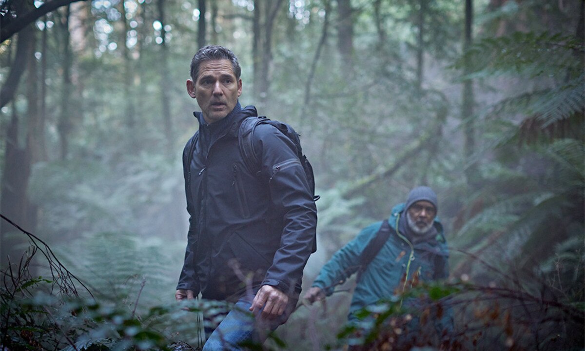 Eric Bana, in character as Aaron Falk, searches in the bush, another man behind him looking in same direction.