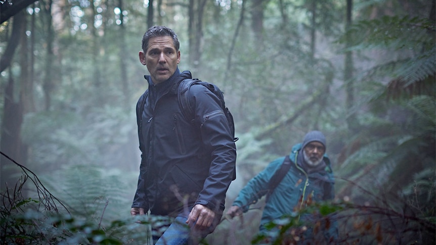 Eric Bana, in character as Aaron Falk, searches in the bush, another man behind him looking in same direction.