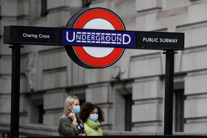 Two women wearing facemasks walk under a sign for the London Underground