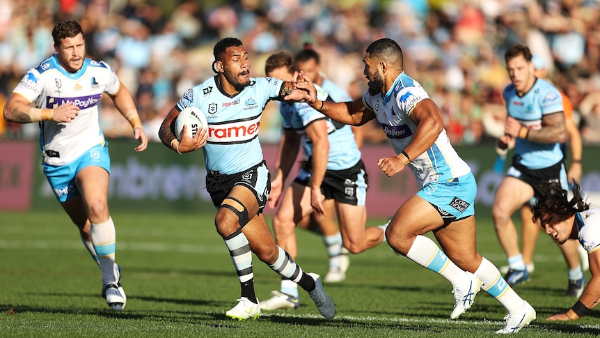 A Cronulla NRL player puts his palm out to keep a defender away as he sprints downfield holding the ball.