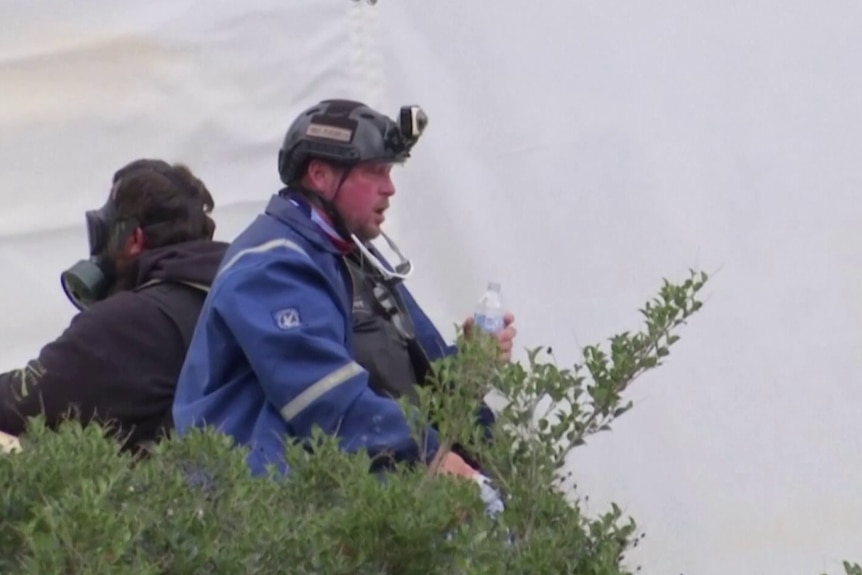 파란색 재킷과 검은색 헬멧을 착용하고 전면에 장착된 카메라를 착용하고 물병을 들고 앉아 있는 남자.