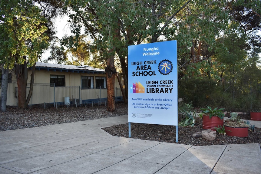 A blue sign that says 'Leigh Creek Area School' in front of a cream-coloured building surrounded by trees.