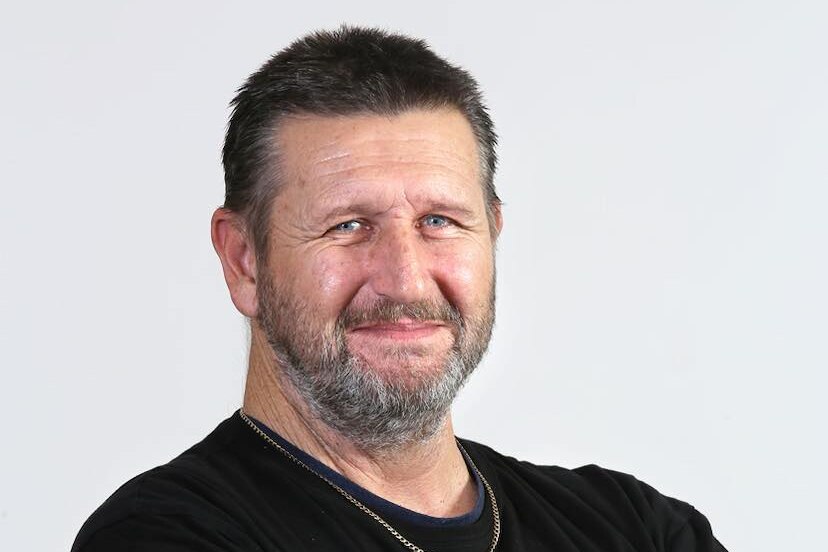 white man with greying hair and beard in chain and black top with arm tattoos smiles at camera with arms crossed