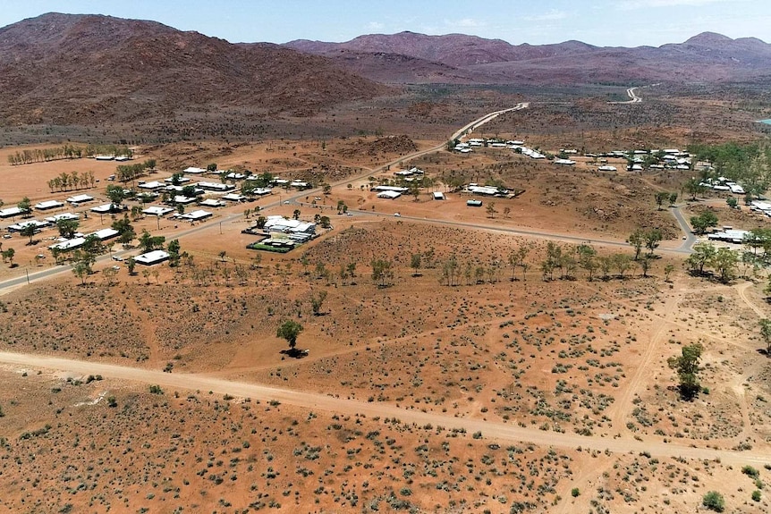 An aerial photo of a remote town in a red arid desert with a few trees and dirt roads against a mountain range.