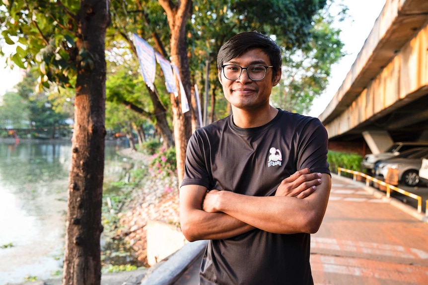 A man with black hair and wearing glasses stands smiling in front of a tree with his arms folded