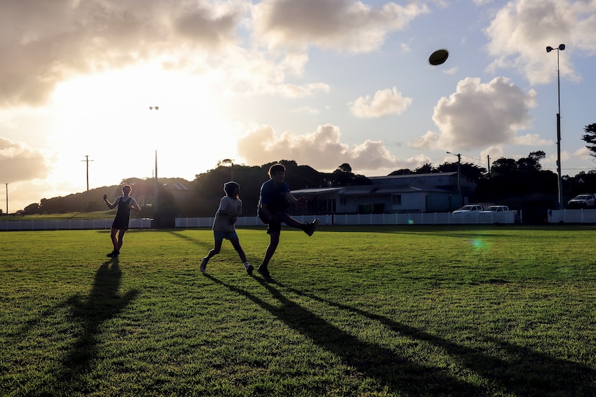 Three kids are silohuetted against the late afternoon sun. They are on a football field kicking a ball