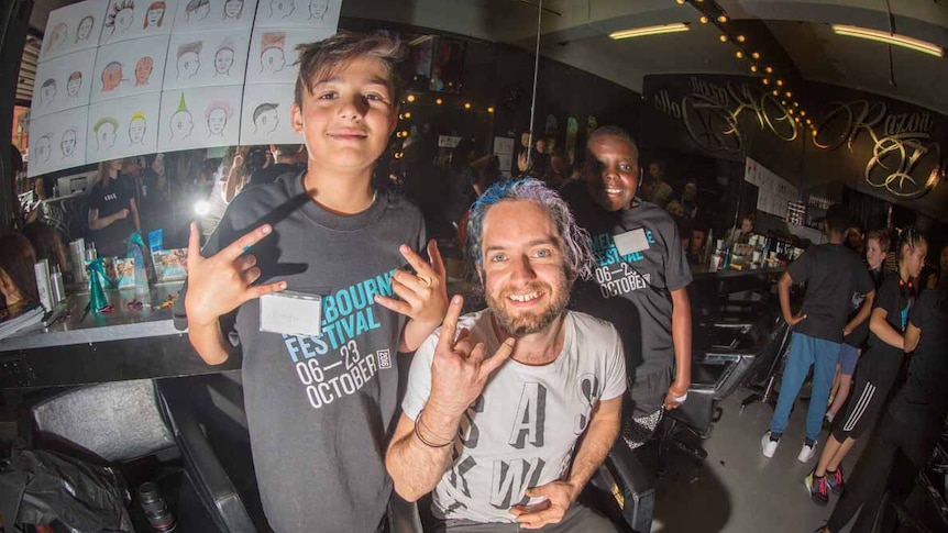 Two schoolboys in hairdressing school pose with their client, who now has blue and purple hair.