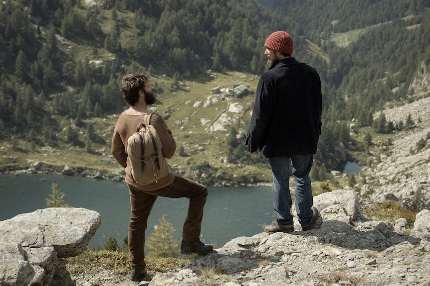 Two Italian men in their 30s, one with dark hair and the other with light hair, stand overlooking a lake in the Italian Alps.