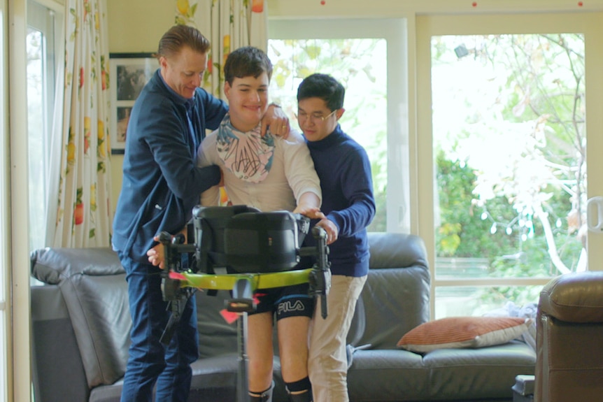 Two men assisting a young man in stand up while holding onto a walking machine