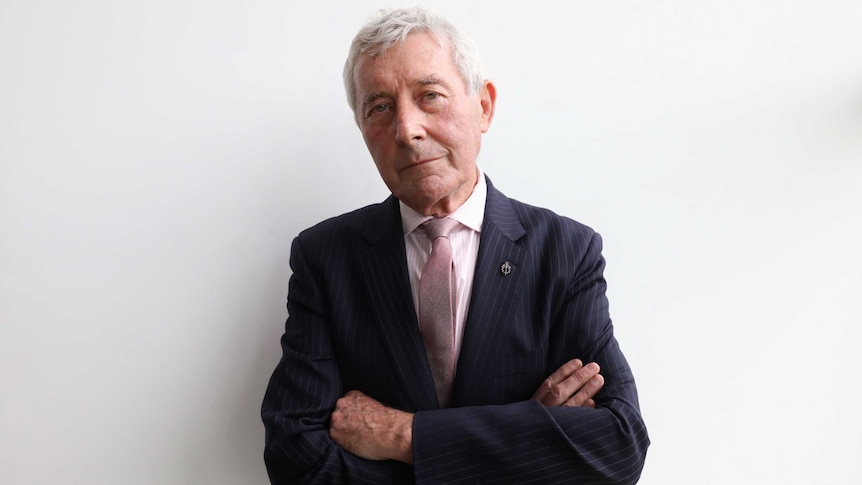 Bernard Collaery looks directly at the camera, with his arms crossed. He's wearing a navy blue suit and pink tie.