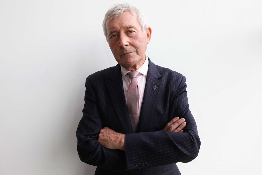 Bernard Collaery looks directly at the camera, with his arms crossed. He's wearing a navy blue suit and pink tie.