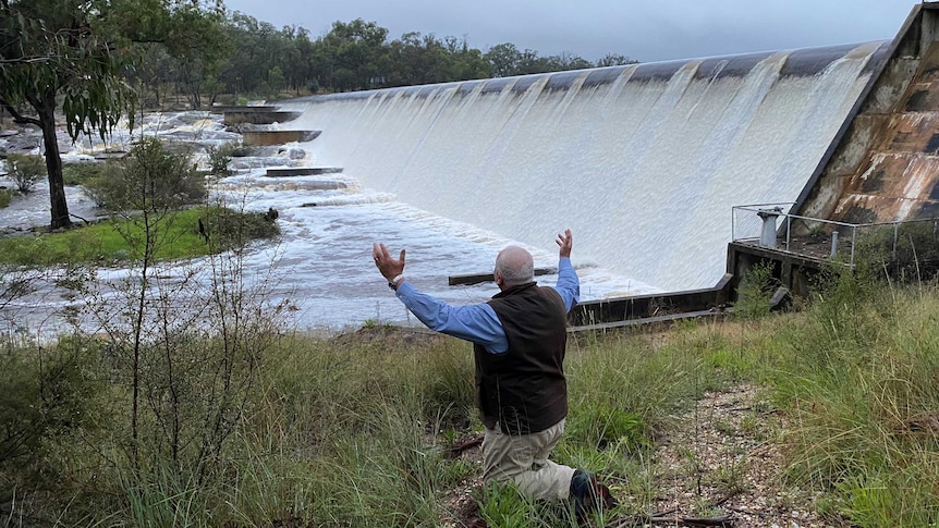 A man on his knees with his arms in the air, looking at water spilling from a dam.