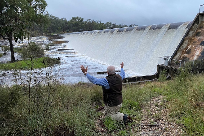 A man on his knees with his arms in the air, looking at water spilling from a dam.