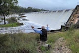 A man on his knees with his arms in the air, looking at water spilling from a dam.