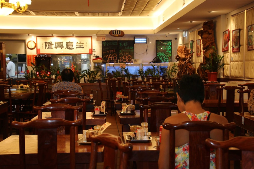 Two diners sit at tables in the restaurant. The wall is decorated with picture menus and specials on the blackboard.