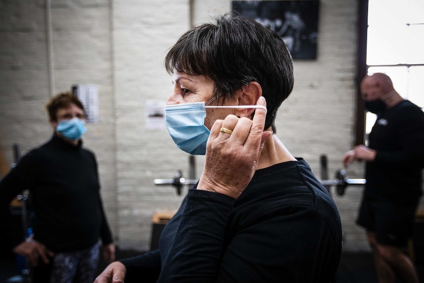 A women viewed in profile puts on a face mask. Two other women watch