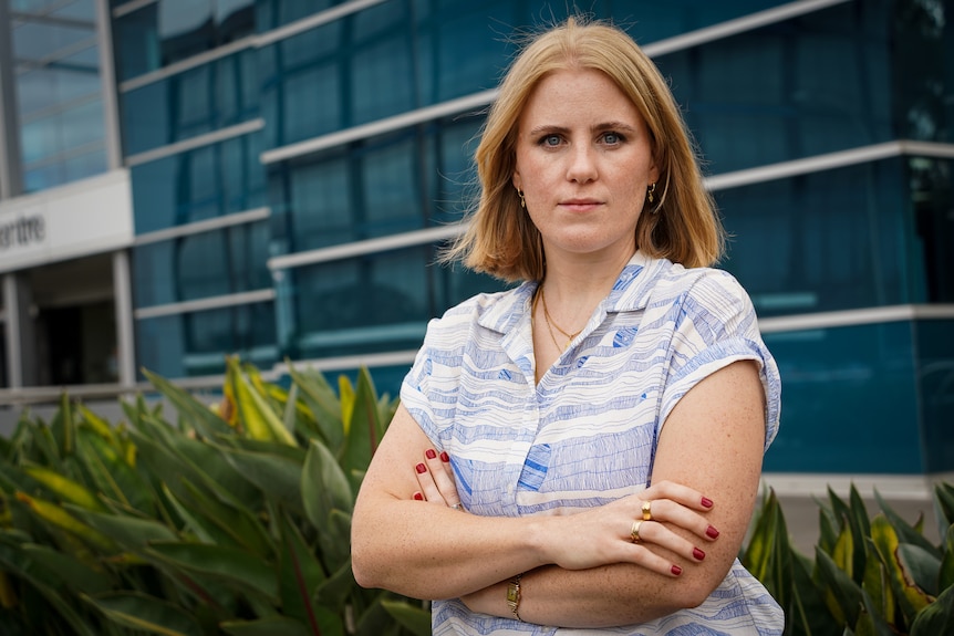 A woman with red hair stands in front of an office building with her arms folded