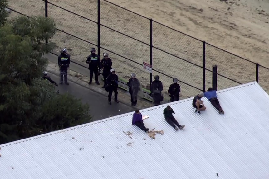 A group of detainees sit and lie on the roof of a building at Banksia Hill Detention Centre, with armed police on the ground.