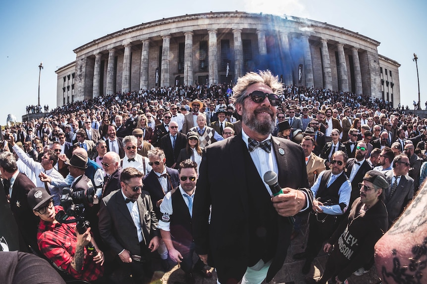 A crowd of several hundred men suits crowd around a man speaking with a microphone