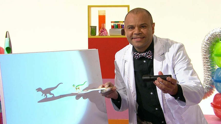 Luke on the Play School Science Time set wearing a lab coat holding a torch and a toy dinosaur making a shadow