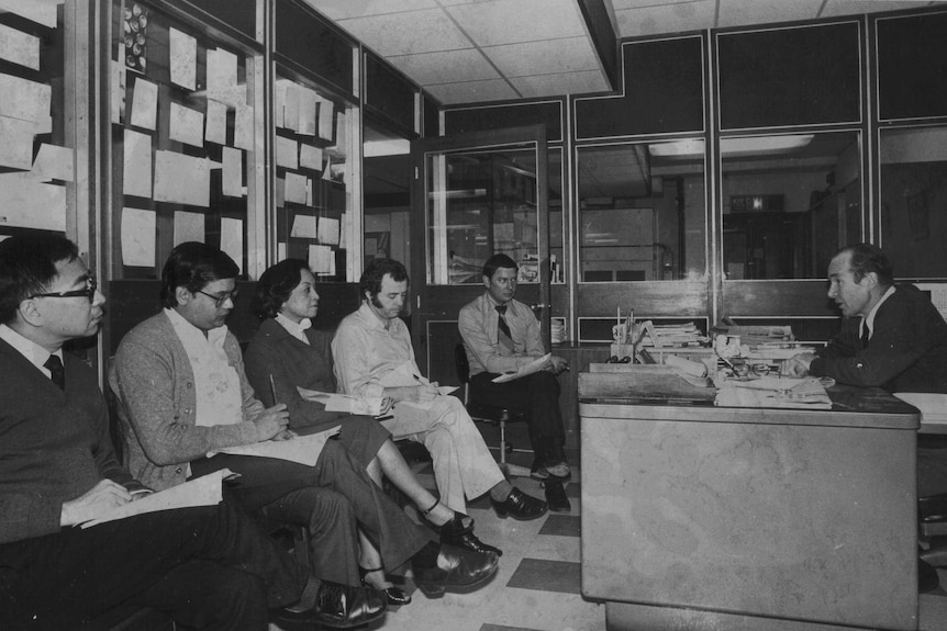Black and white photo of Hutcheon sitting behind a desk talking to journalists holding notepads and pens.