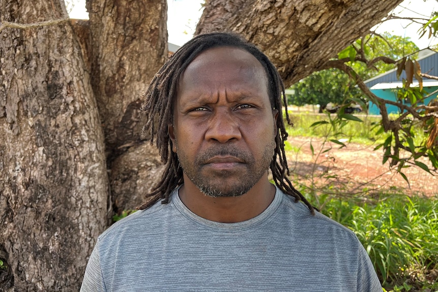 Indigenous man with dreadlocks stands in front of tree and looks at camera
