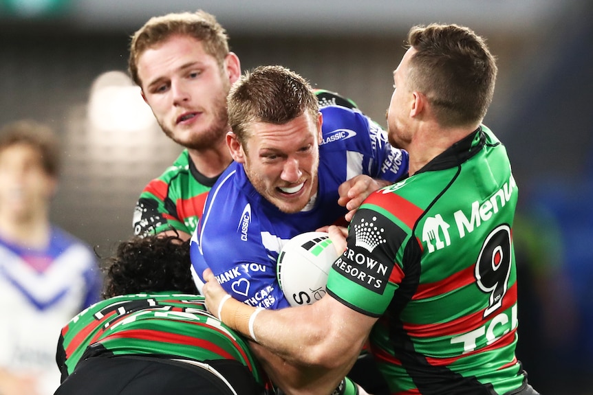 A Canterbury NRL player is tackled by three South Sydney opponents.