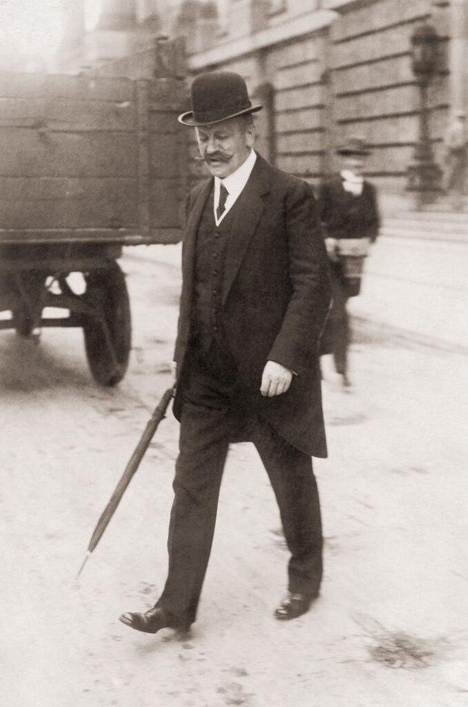 Black and white photograph of an older man with a big mustache crossing the road with a bowler hat and a cane.