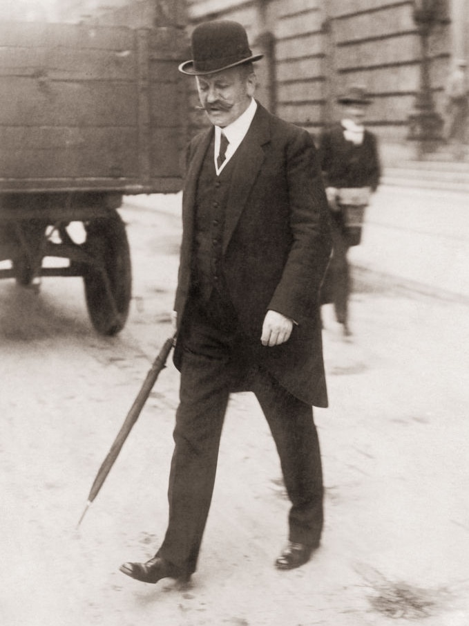 Black and white photograph of an older man with a big mustache crossing the road with a bowler hat and a cane.